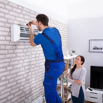 Smiling Woman Looking At Male Technician Repairing Air Conditioner With Screwdriver In Home